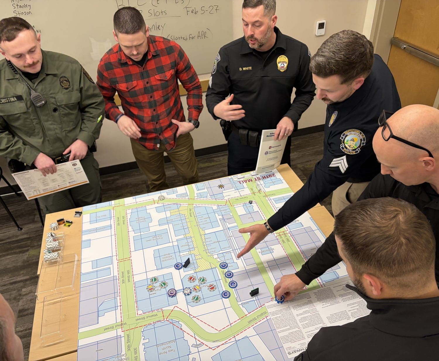 Law enforcement, fire, and EMS responders gathered around a Counterstrike Basic environment map, an aerial street map of a real location, one pointing at a position with resource chips staged on the table