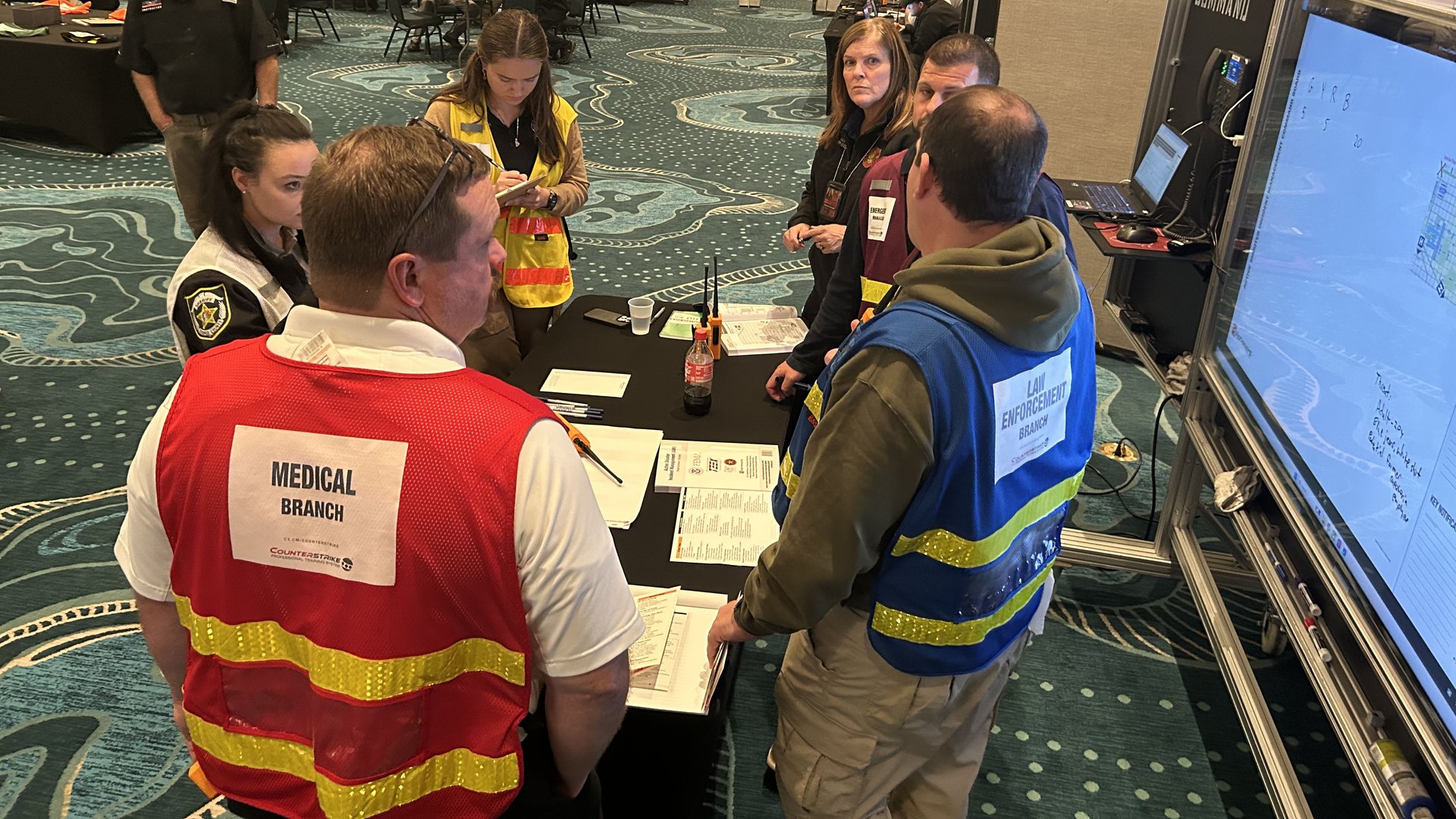 Law Enforcement Branch and Medical Branch Directors in labeled vests confer with command staff at the Command Post during an active shooter training exercise