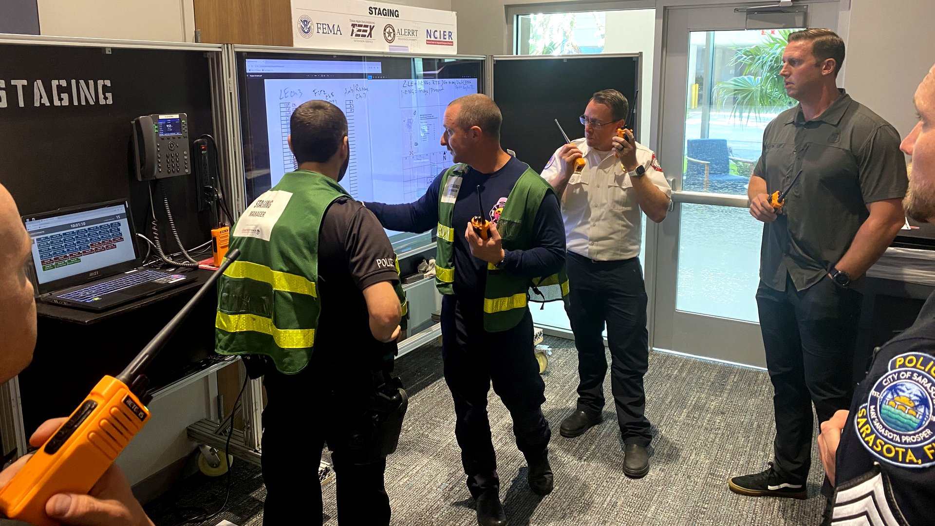 Staging Manager in a green vest briefs police and fire responders at a staging board during an active shooter incident management course